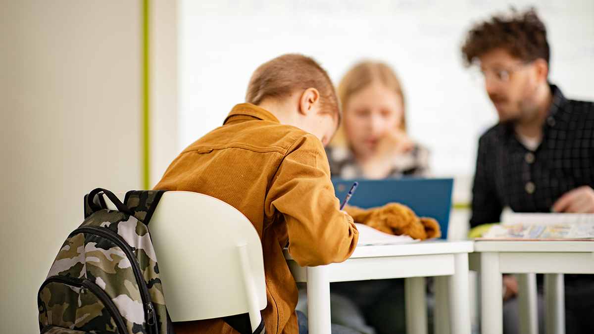 Opettaja ja kaksi oppilasta istuvat pulpettien ääressä opiskelemassa.