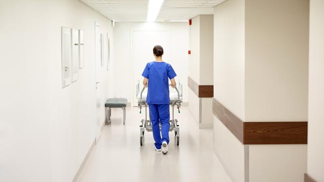 Kuvassa hoitaja työntää sairaalasänkyä sairaalan käytävällä. Hänellä on selkä kameraan päin.