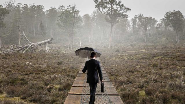 Tummaan pukuun pukeutunut mies kävelee metsässä sateenvarjon kanssa. Kuva: Pixhill