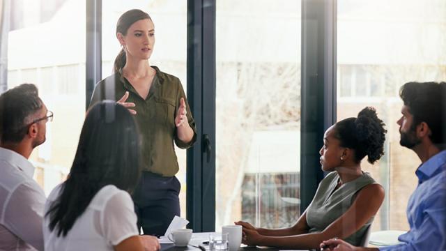 Joukko kollegoita istuu pöydän ääressä ja kuuntelee, kun yksi heistä puhuu seisaaltaan.
