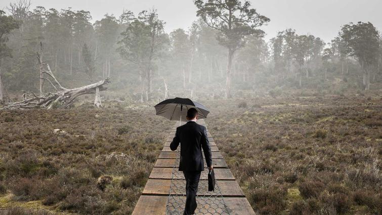 Tummaan pukuun  pukeutunut mies kävelee metsässä sateenvarjon kanssa. Kuva: Pixhill
