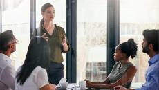 Joukko kollegoita istuu pöydän ääressä ja kuuntelee, kun yksi heistä puhuu seisaaltaan.